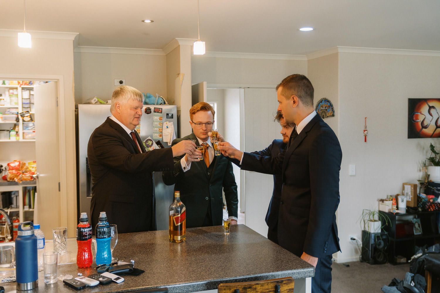 Three men in suits toasting with whiskey glasses in a modern kitchen during a wedding celebration.