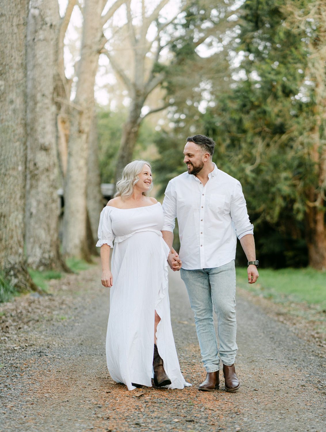 Couple walking hand in hand on a tree-lined gravel path, wearing white outfits, smiling at each other.