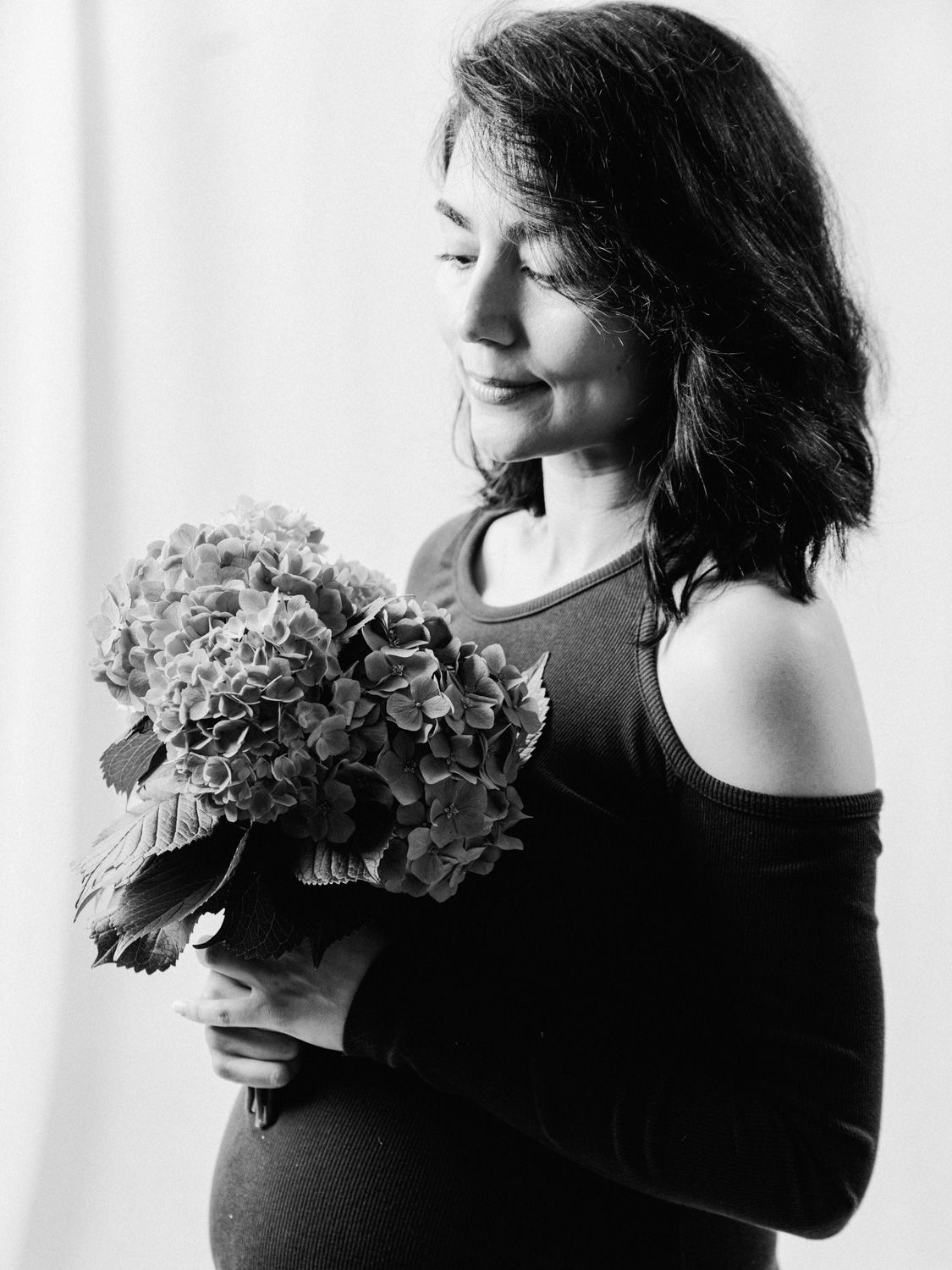 Black and white maternity photo of a woman holding hydrangea flowers, smiling softly with eyes closed.