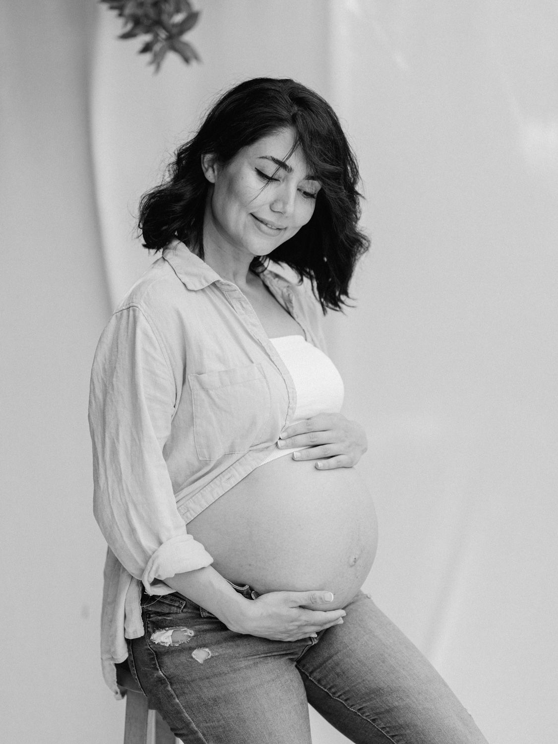 Black and white maternity photo of a pregnant woman in jeans cradling her baby bump with eyes closed.