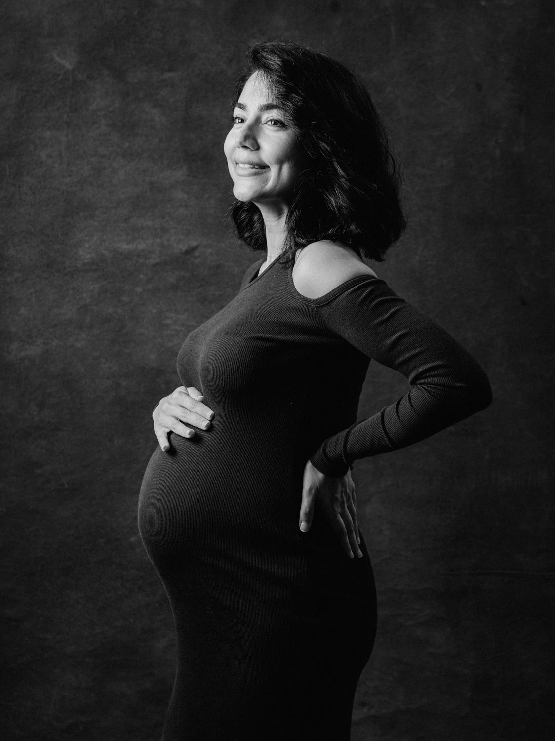 Black and white maternity portrait of a smiling woman in a form-fitting dress cradling her baby bump.