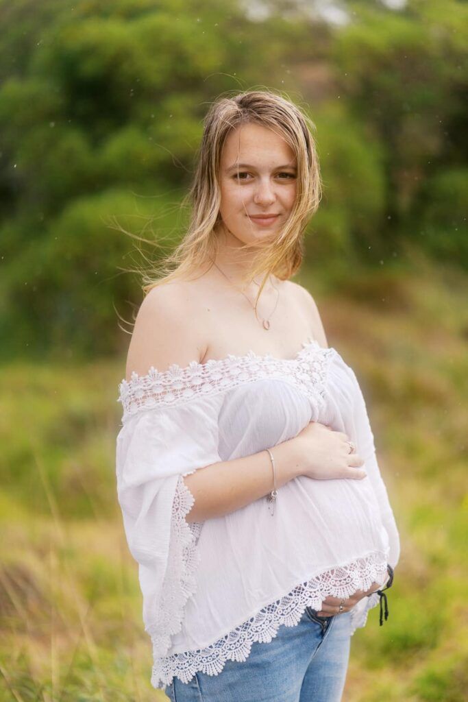 Pregnant woman in white off-shoulder top and jeans poses outdoors in a green field with wind-blown hair.