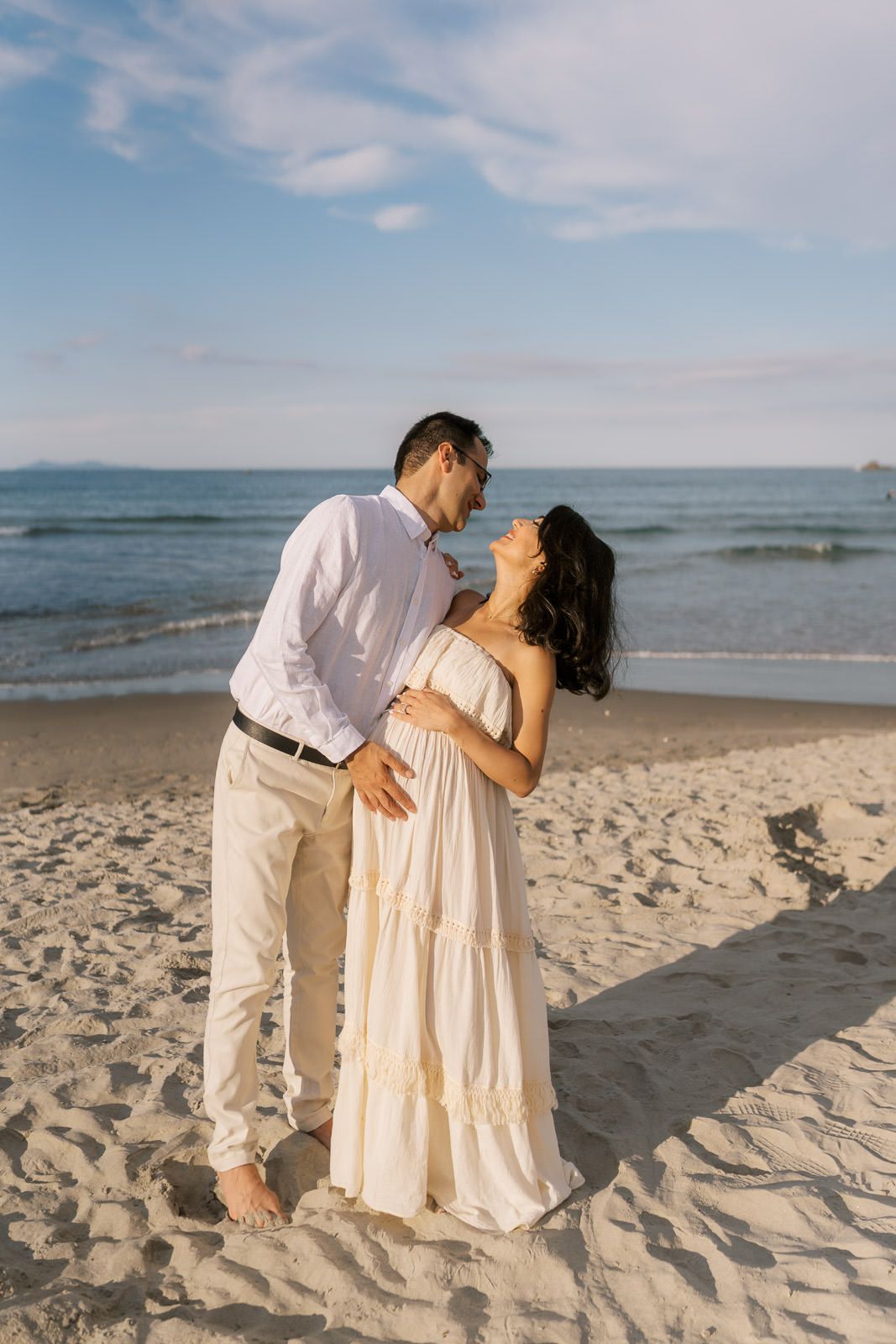 A couple embraces romantically on a sandy beach, the woman in a white dress leaning back as waves roll in behind them.