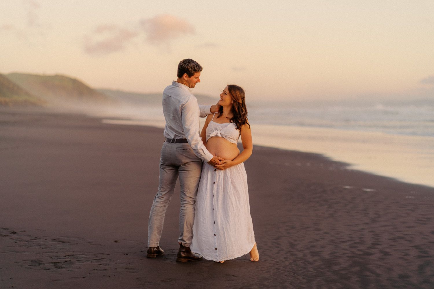 Couple sharing a tender moment on a black sand beach at sunset, woman in white maternity dress cradling her baby bump.