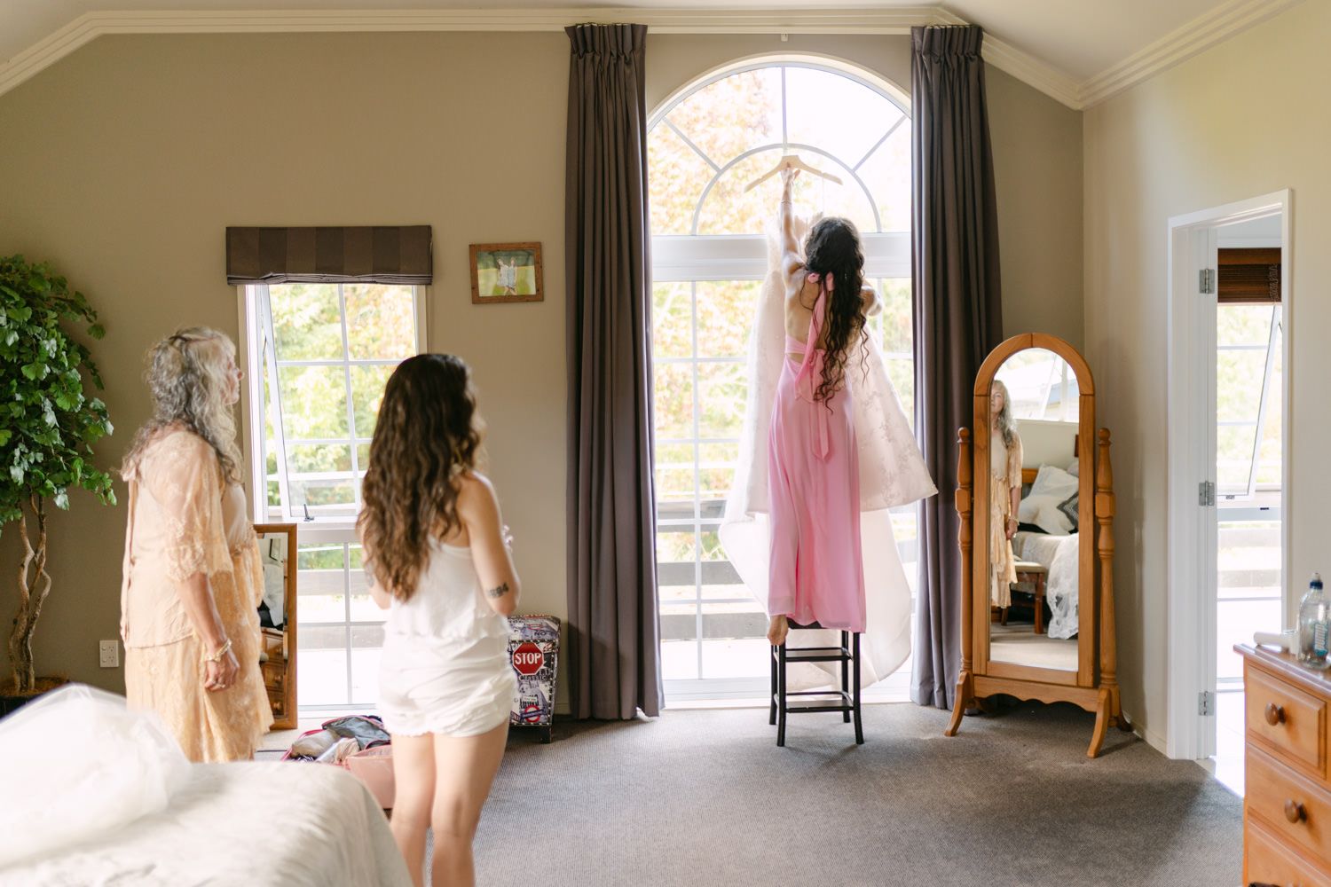 Bride and bridesmaids in robes looking out large window with floor mirror in elegant bedroom on wedding day.