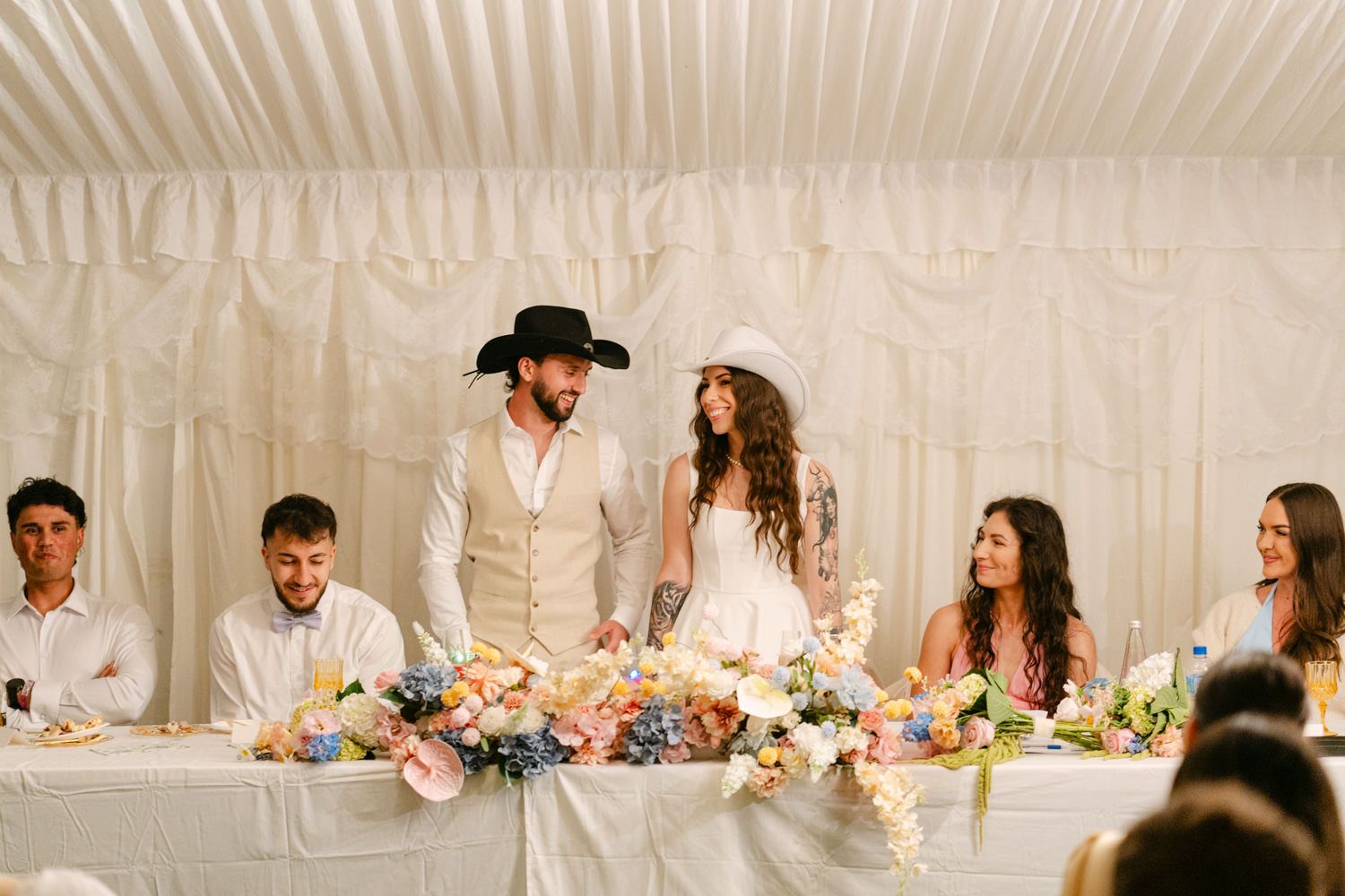 Bride and groom stand at floral-adorned wedding reception table in white tent, surrounded by smiling guests.
