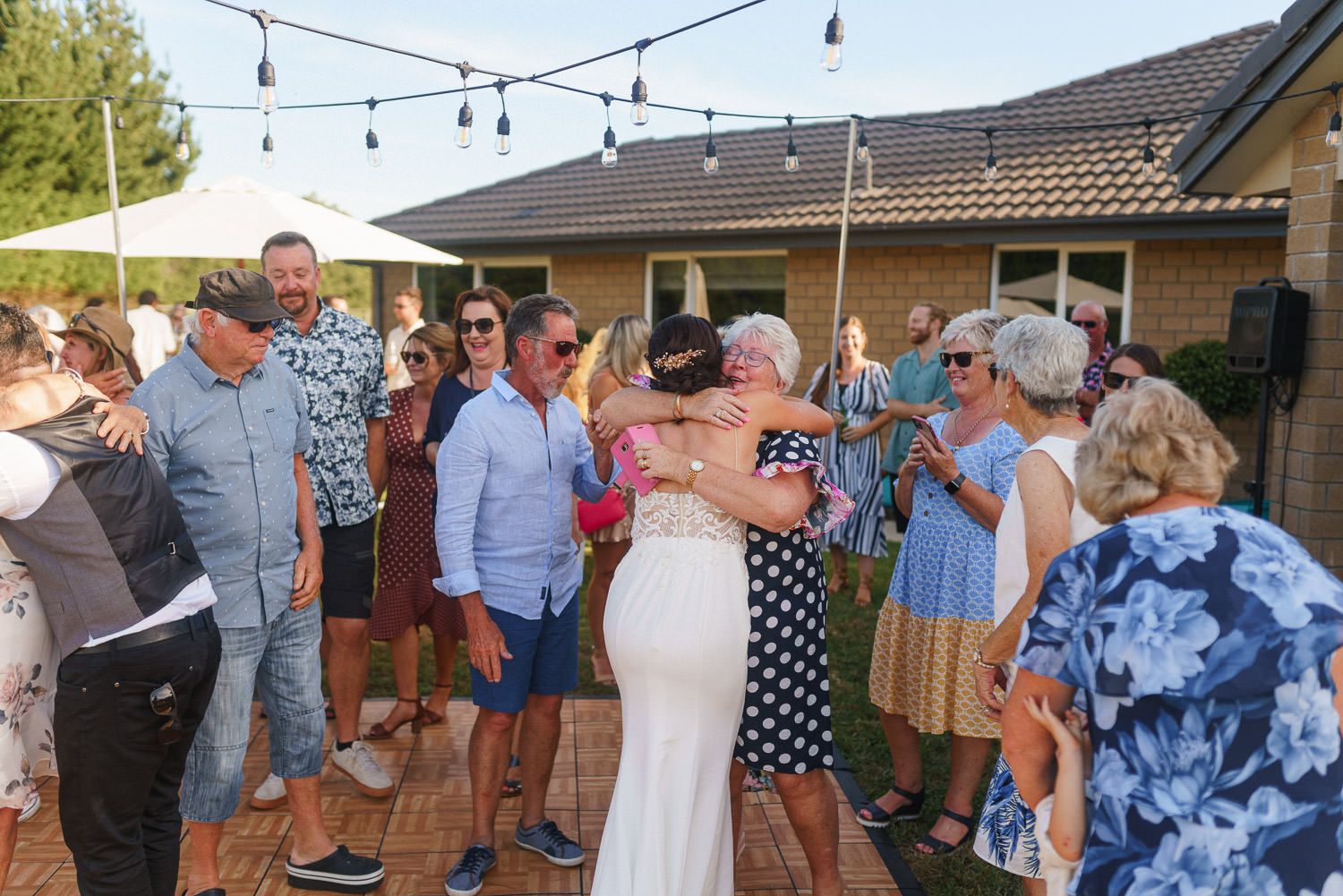 Bride in white gown hugs guest surrounded by smiling wedding party outdoors under string lights.
