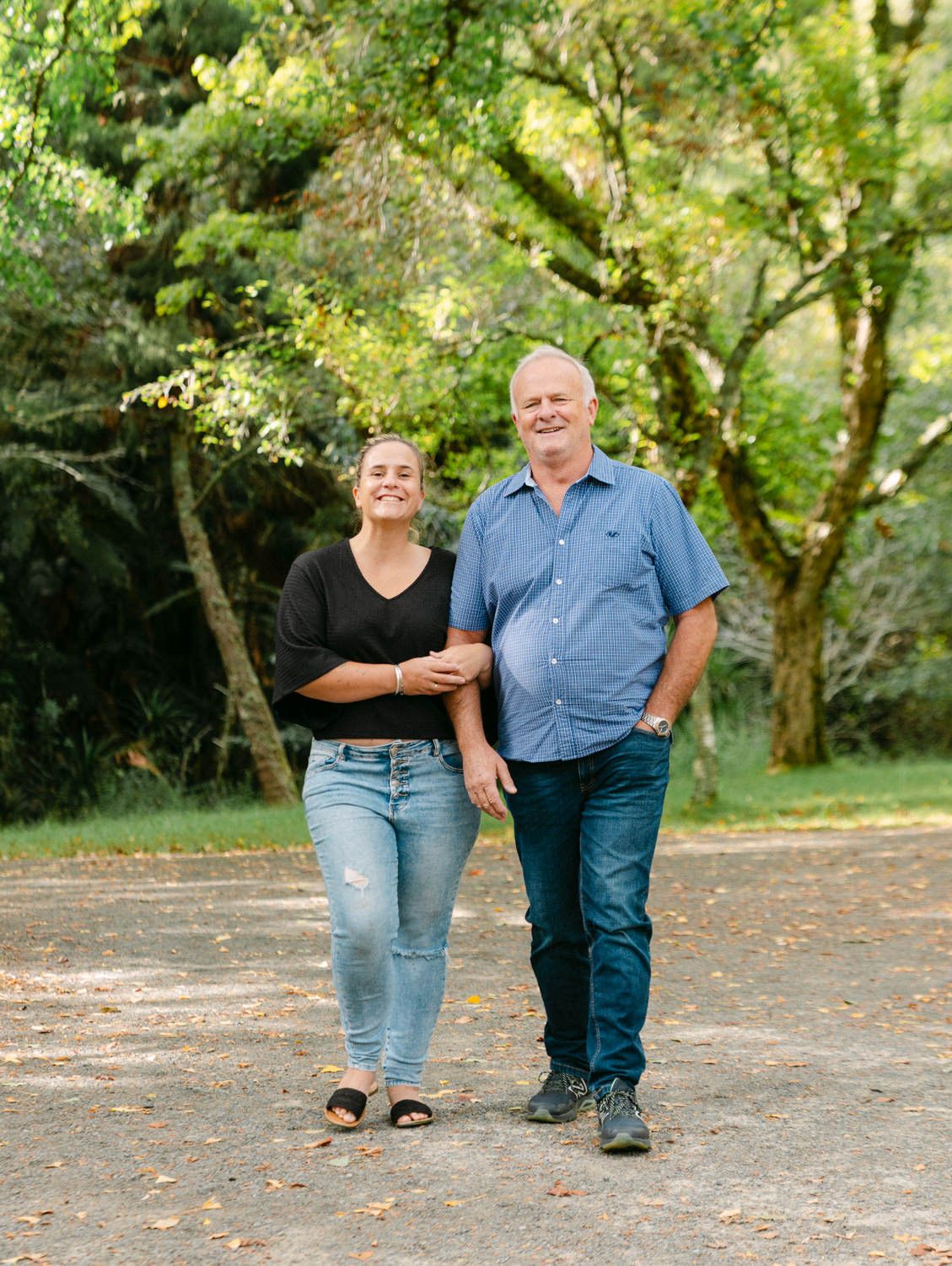 A smiling young woman and older man walk together on a gravel path surrounded by lush green trees.
