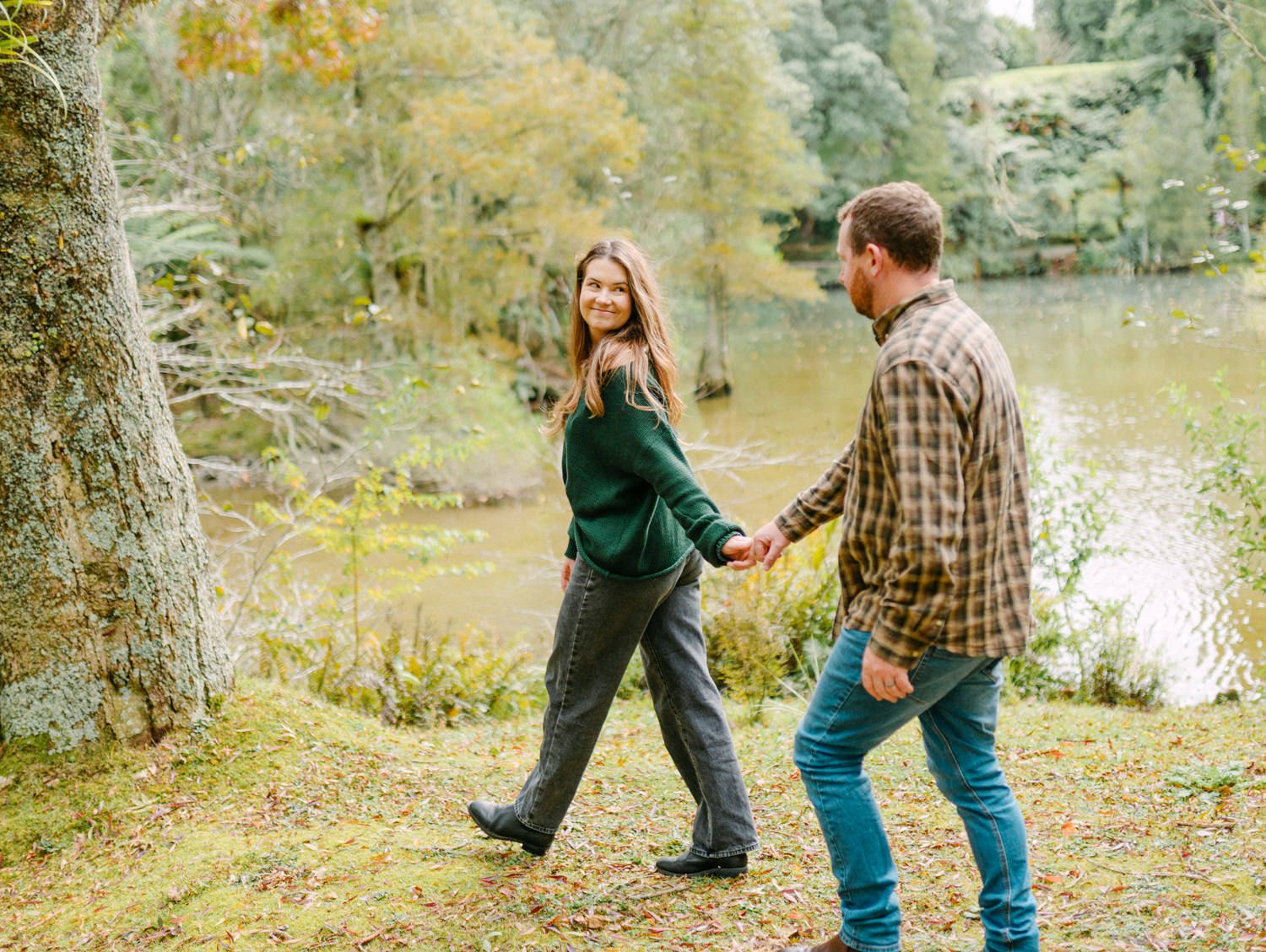 Couple holding hands walking along a scenic autumn lakeside path surrounded by colorful fall foliage.