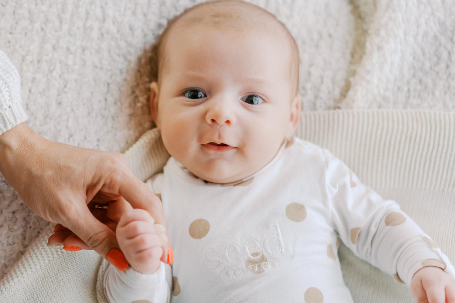 Adorable baby in polka dot onesie lying on white blanket, holding an adult's hand with bright blue eyes.