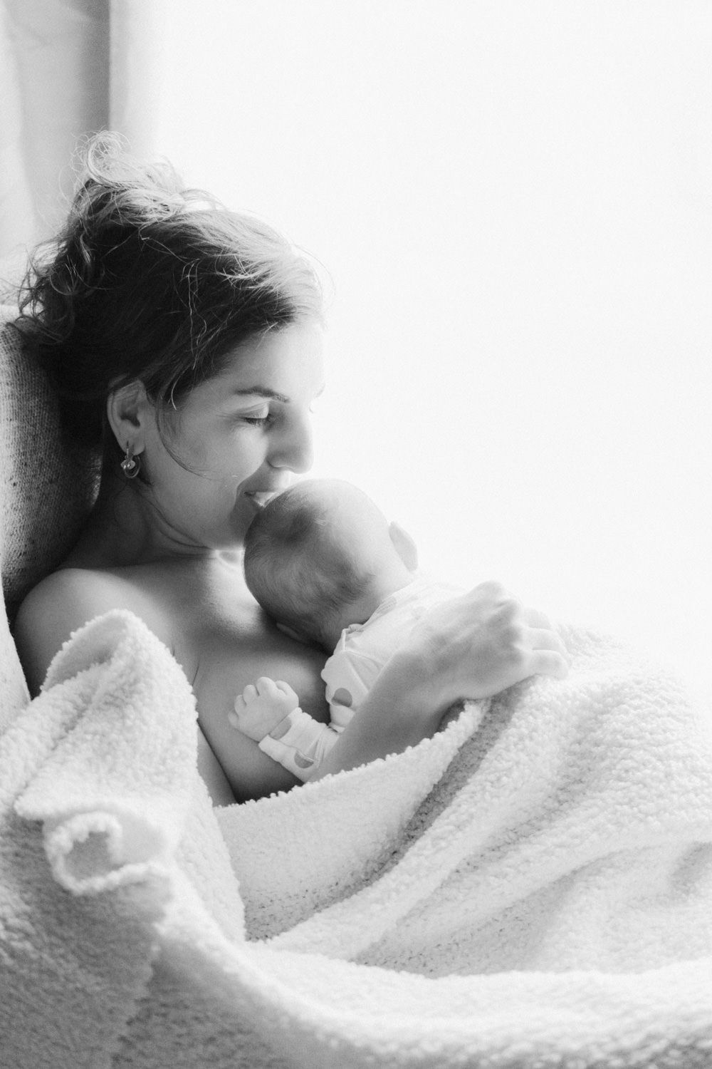 Black and white photo of a mother tenderly kissing her newborn baby wrapped in a soft white blanket.