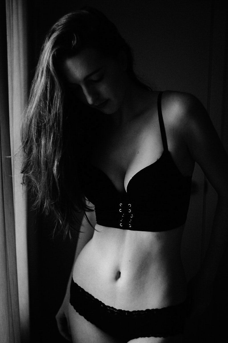 Black and white boudoir photo of a young woman in dark lingerie posing by a window in dramatic lighting.