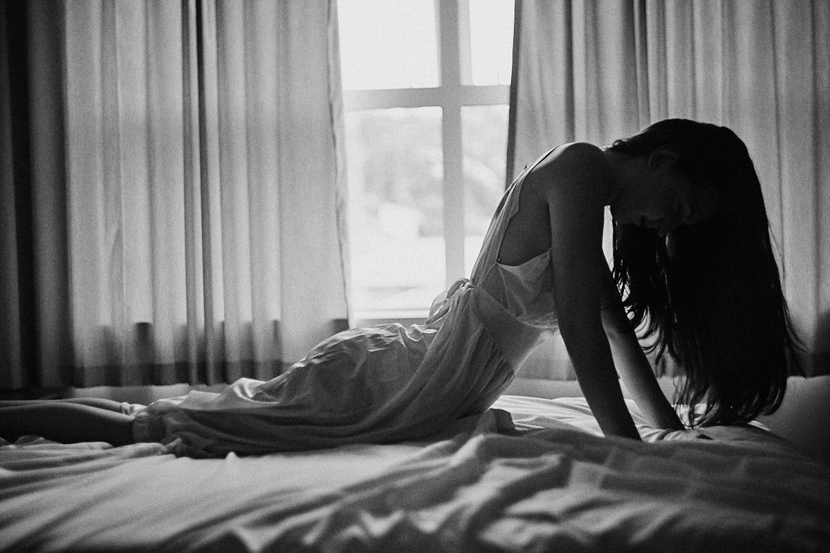 Black and white photo of a woman in a white dress sitting on a bed near a window, head bowed in contemplation.