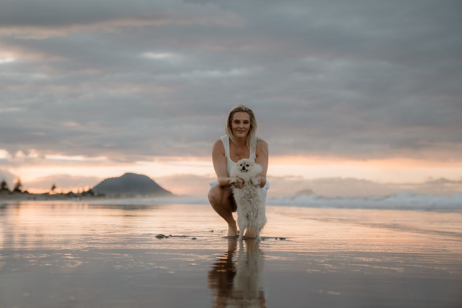 Woman crouching on a reflective beach at sunset, holding a small white dog, with a misty mountain in the background.