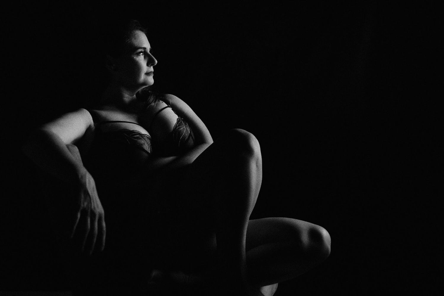 Black and white portrait of a woman sitting in a chair, gazing thoughtfully in dramatic low-key lighting.