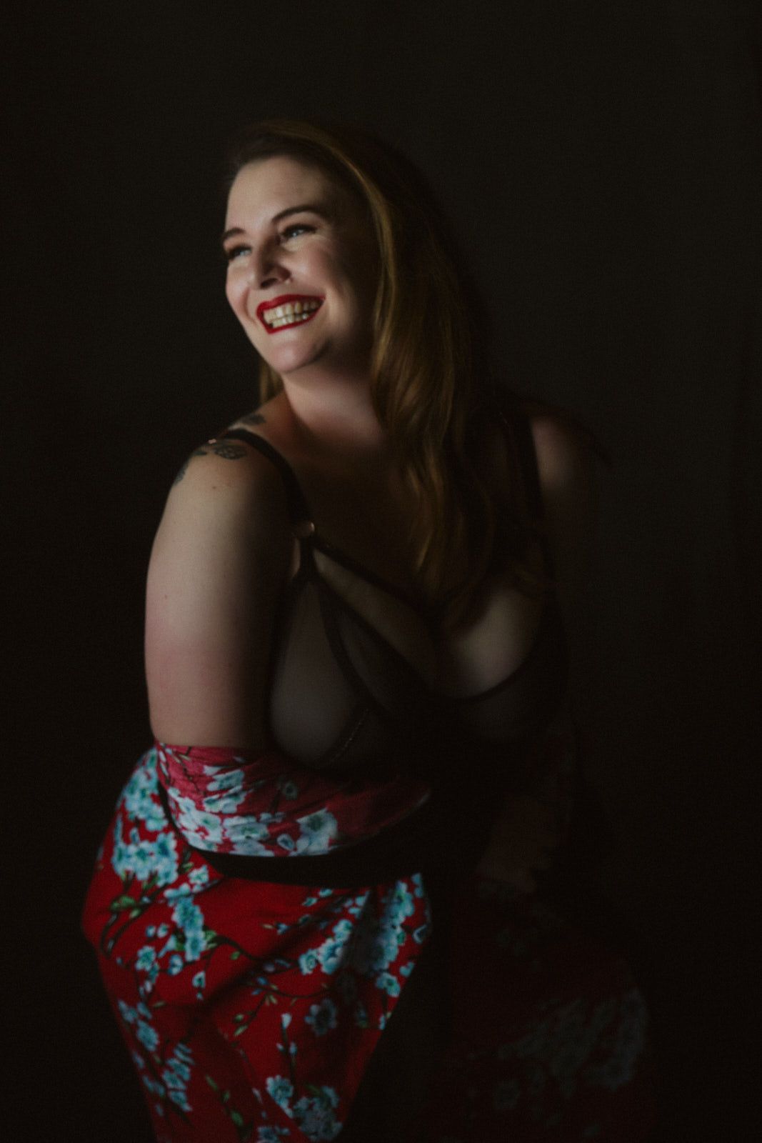 Smiling woman in black bralette and red floral skirt poses against dark background with red lipstick.