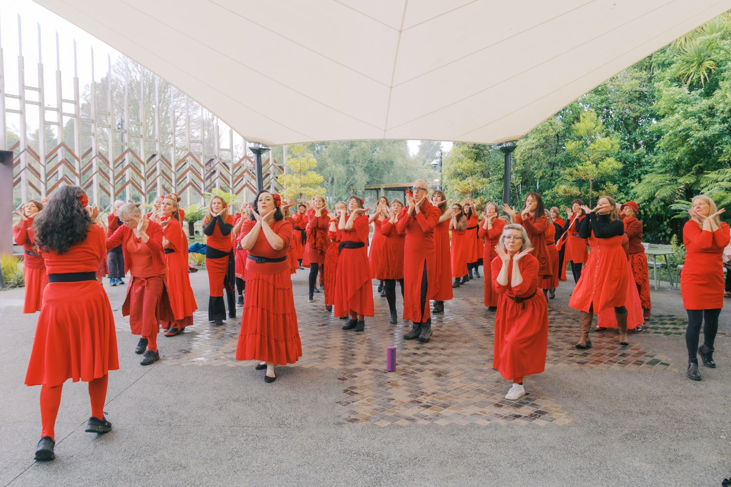Large group of women in matching red dresses performing a synchronized outdoor dance under a white canopy.