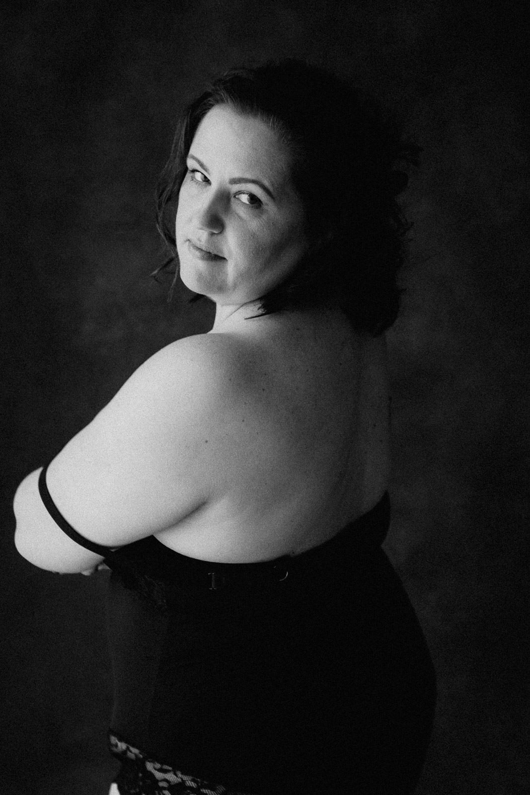 Black and white portrait of a woman in a strapless black dress, looking over her shoulder against a dark background.