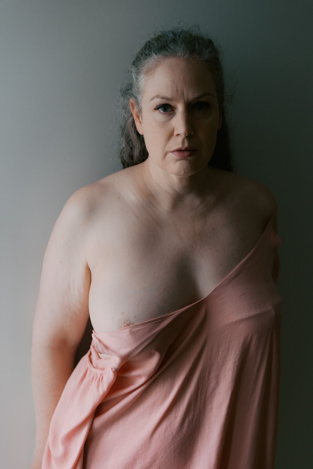 Mature woman with gray hair wearing a pink strapless dress, posing against a gray background.