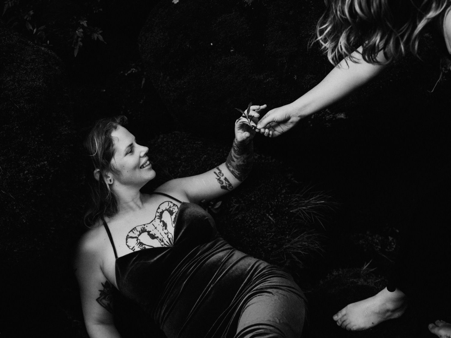 Black and white photo of a woman lying on grass, smiling up while holding hands with another person standing above her.
