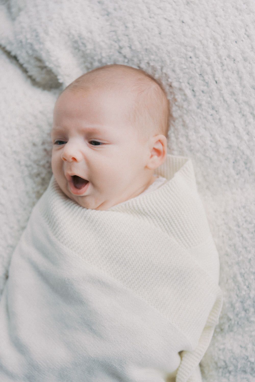 A newborn baby swaddled in a cream knit blanket lying on a soft white surface with mouth open.