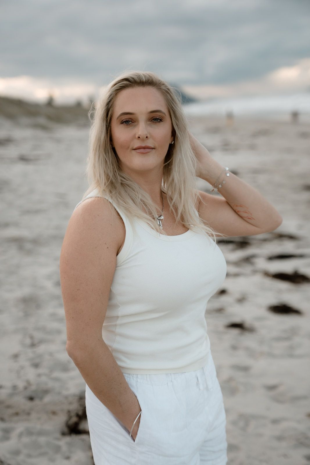 Blonde woman in white tank top and pants poses on a sandy beach with cloudy skies behind her.