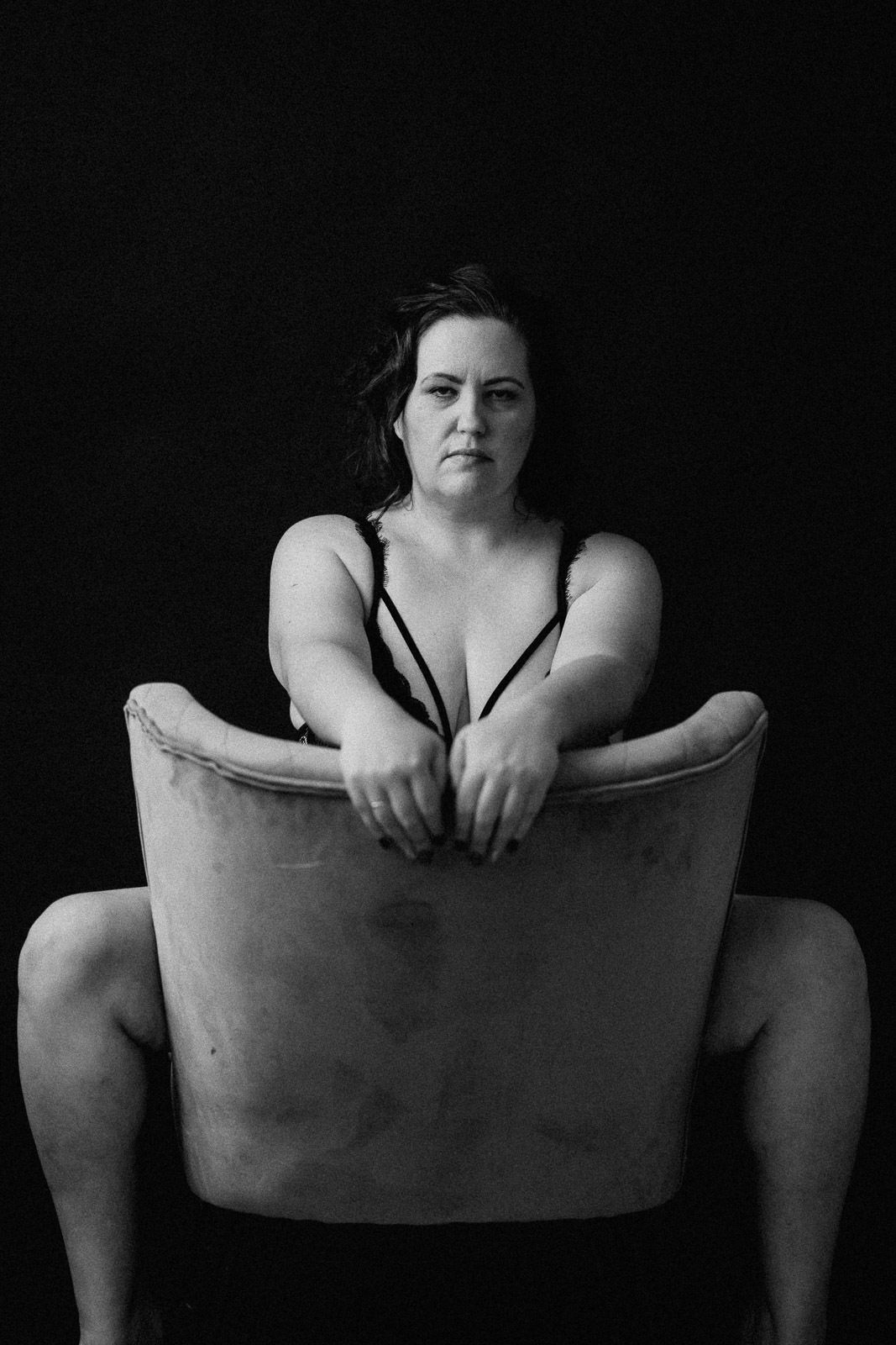 Black and white boudoir portrait of a woman in lingerie posing behind a vintage armchair on dark background.