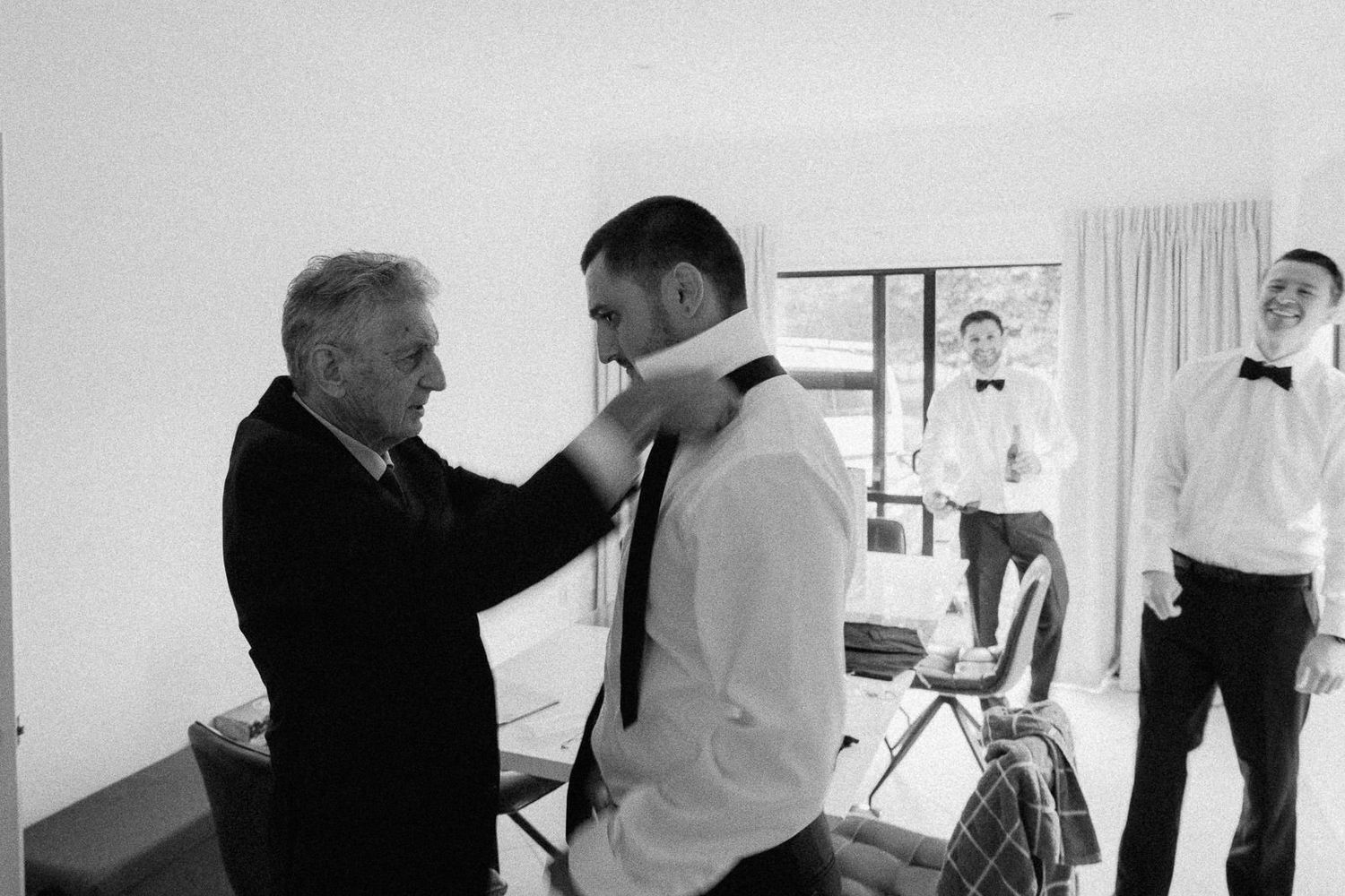 Black and white photo of an older man helping a groom with his tie before a wedding ceremony.