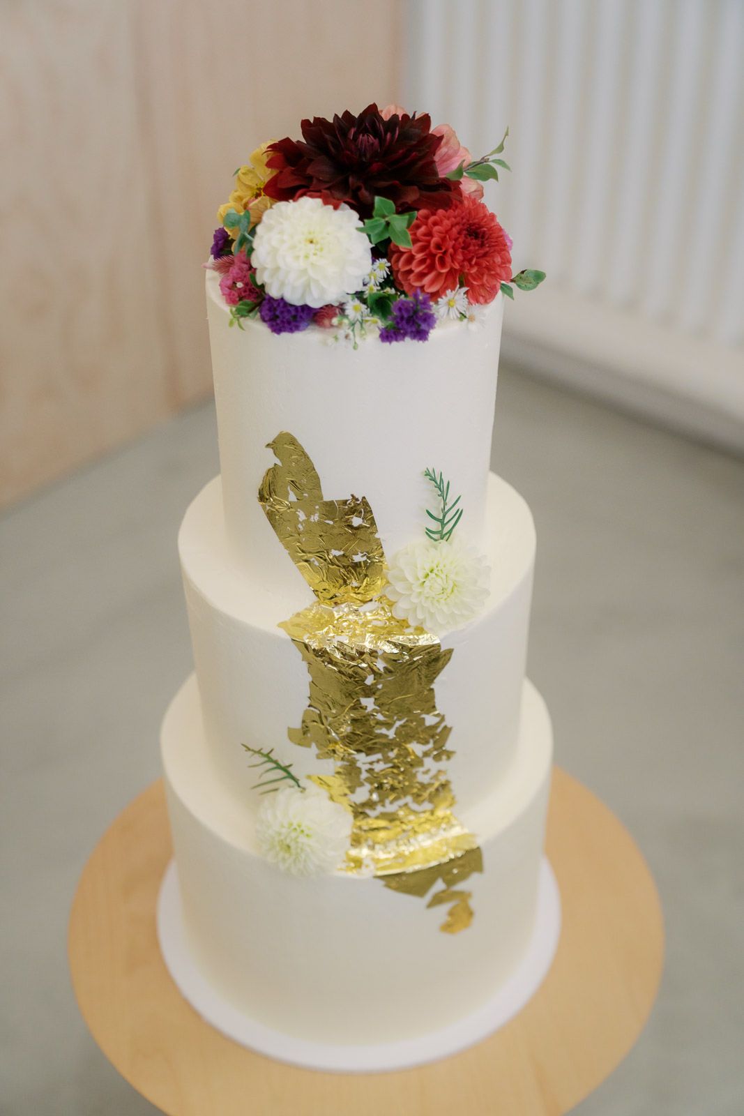 Three-tier white wedding cake with gold leaf detail and colorful fresh flowers on top.