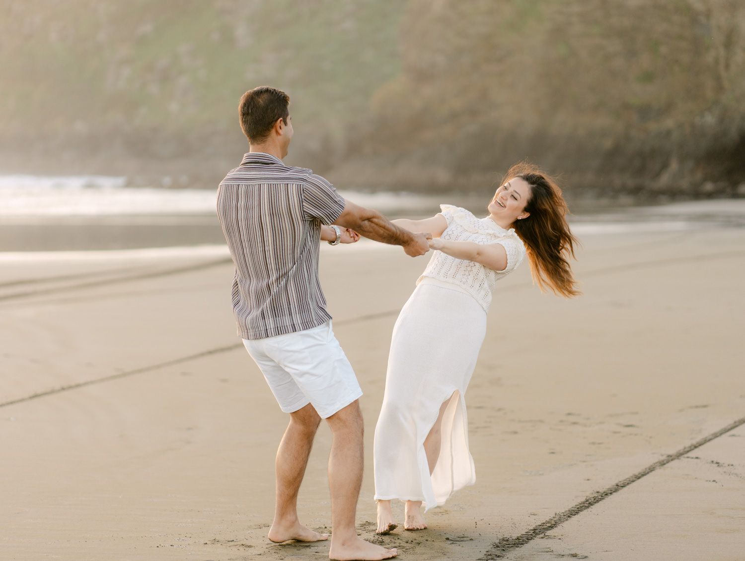A couple dances joyfully on a sandy beach, woman in white dress, man in striped shirt and white shorts.
