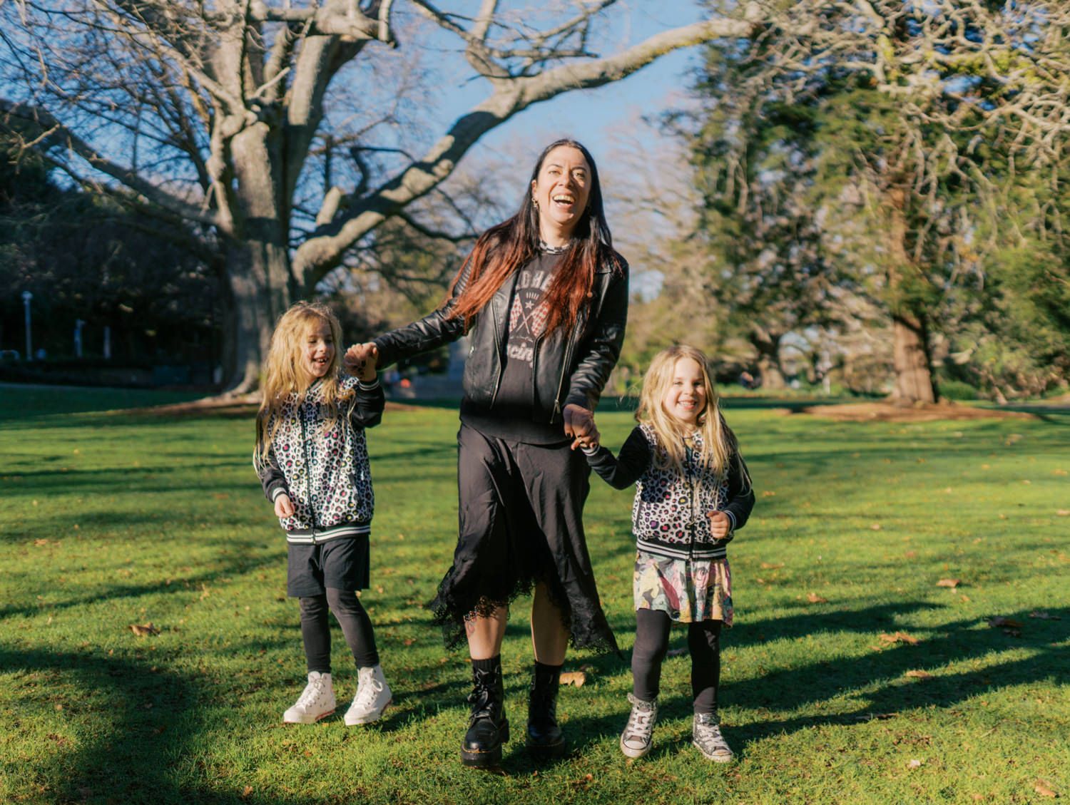 Woman with red hair holds hands with two young girls in matching jackets, laughing in a sunny green park.