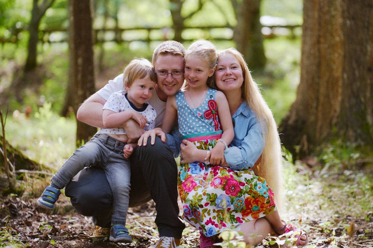 Happy family of four smiling together in a lush green forest setting, parents and two young children posing outdoors.