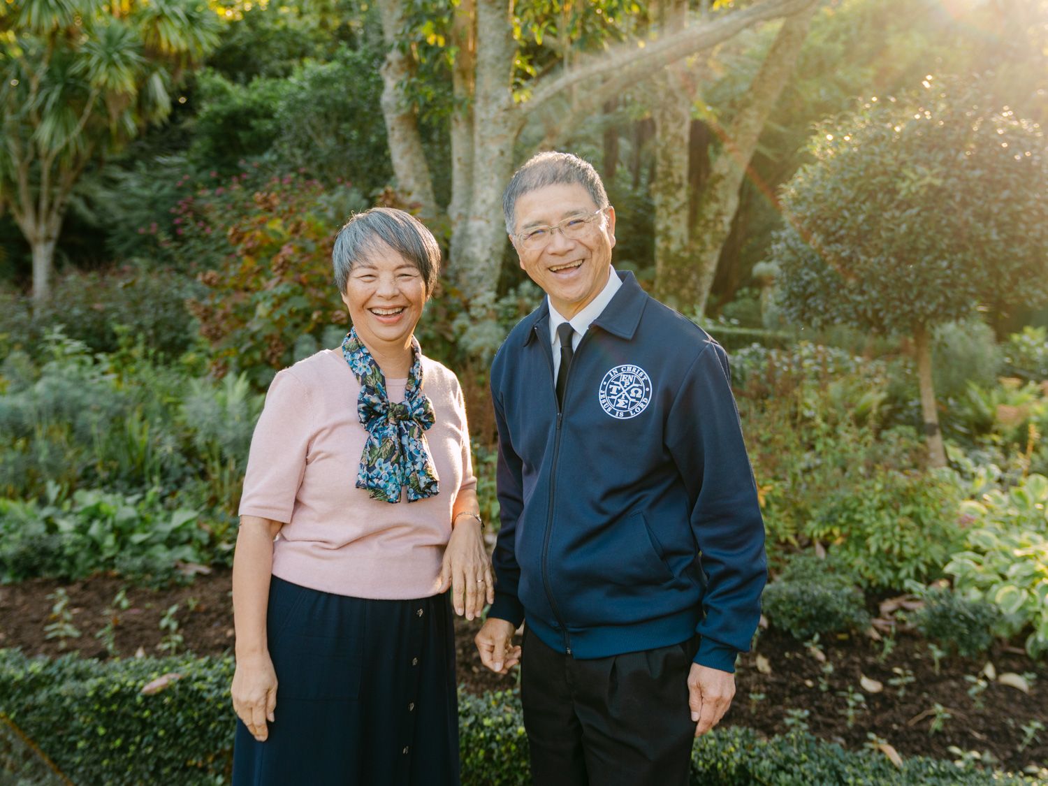 Elderly Asian couple smiling and holding hands outdoors in a lush green garden with warm sunlight.