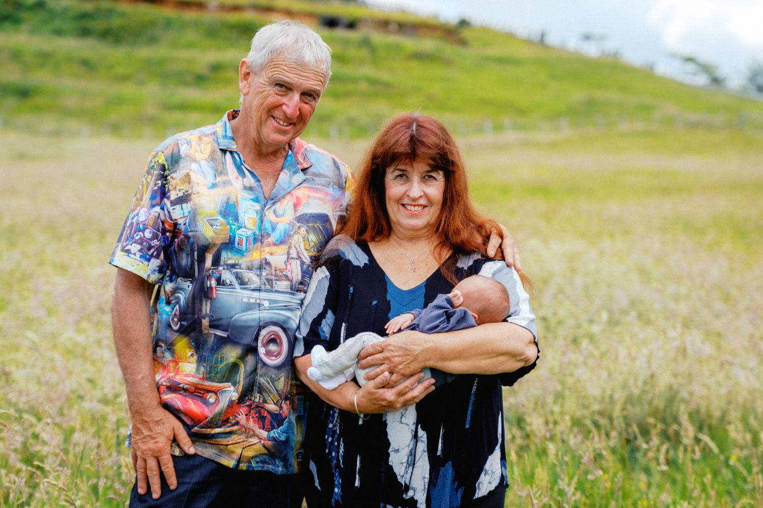 Couple standing in green field holding newborn baby, man in colorful Hawaiian shirt, woman with red hair smiling.