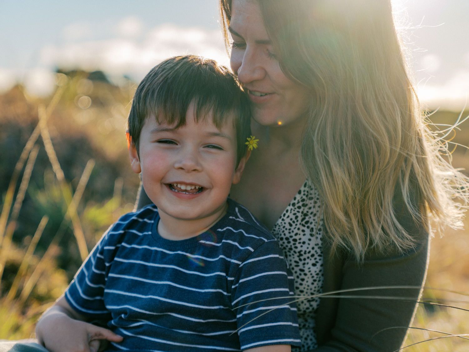 A smiling toddler boy sits with his mother outdoors in a sunny field, both looking happy and relaxed.