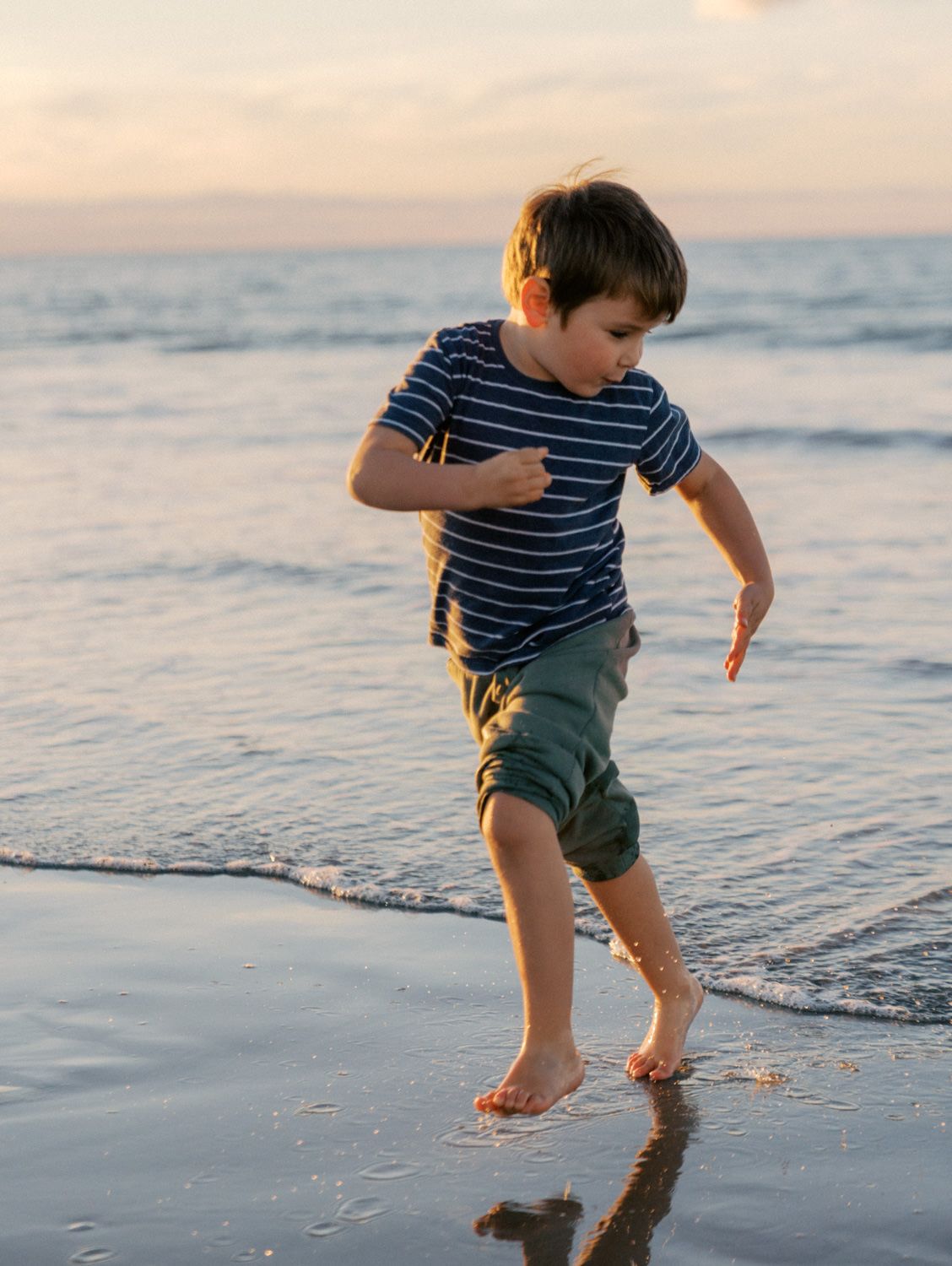 A young boy in a striped shirt and green shorts runs barefoot along the shoreline at sunset.