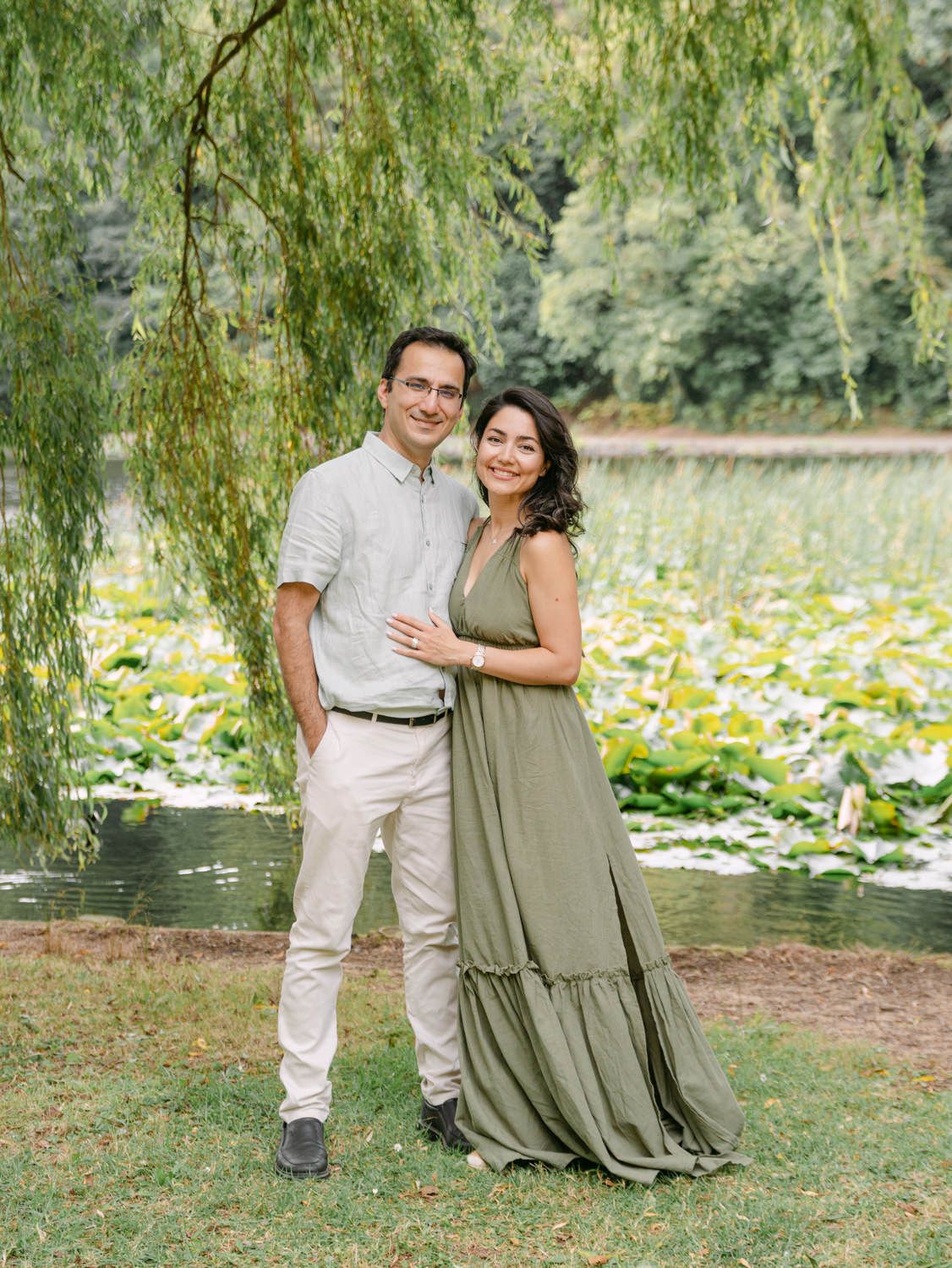 Couple posing together near a serene pond with lily pads and weeping willow trees in a lush green garden.