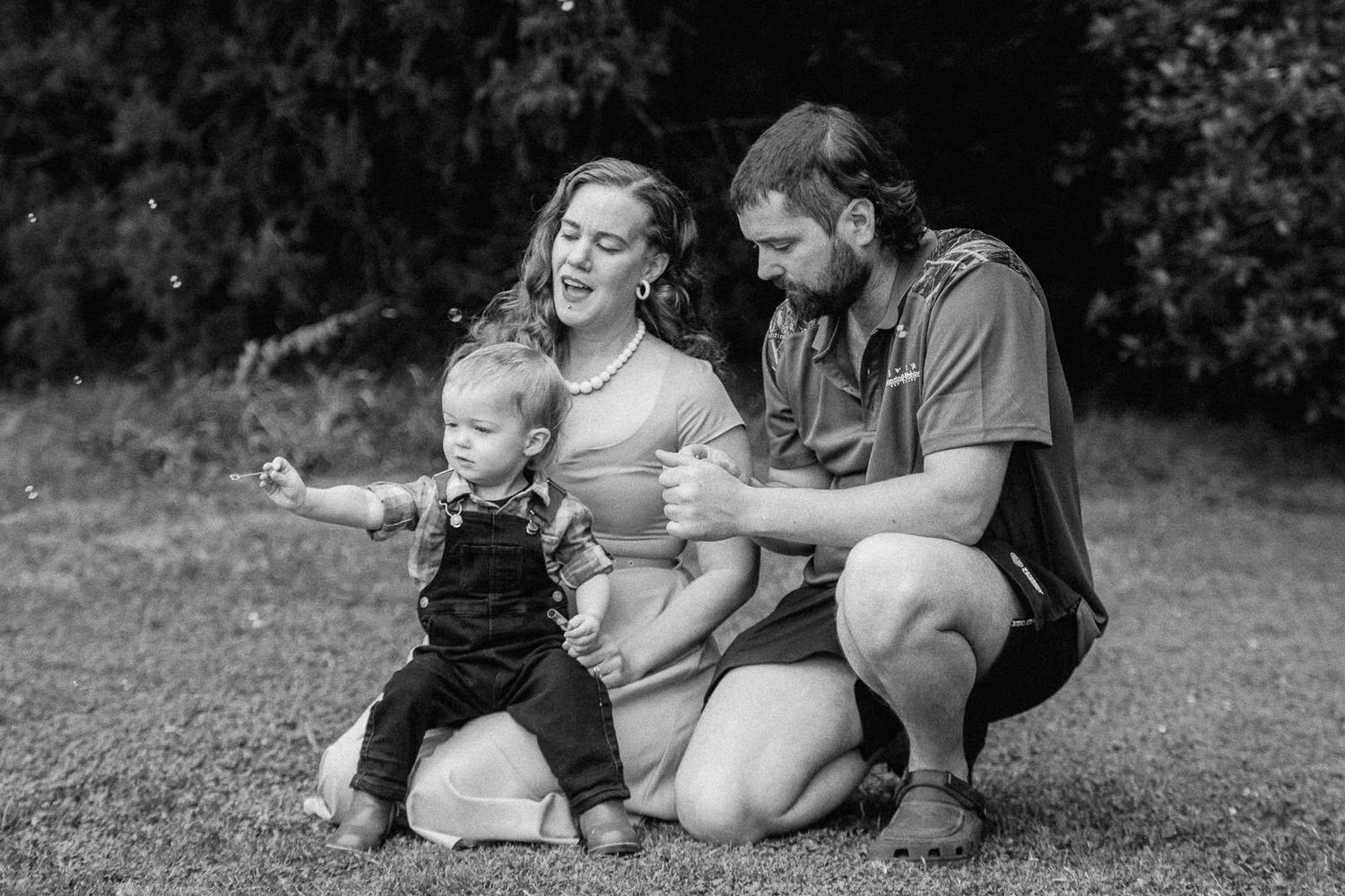 Black and white photo of a young family with a toddler taking first steps on grass outdoors.