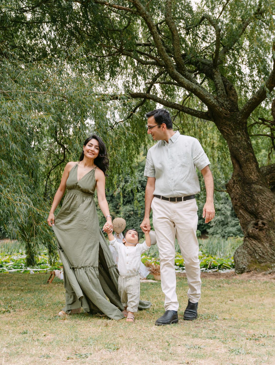 A happy family of three walks hand-in-hand under a willow tree, mom in olive maxi dress, dad in neutral tones.