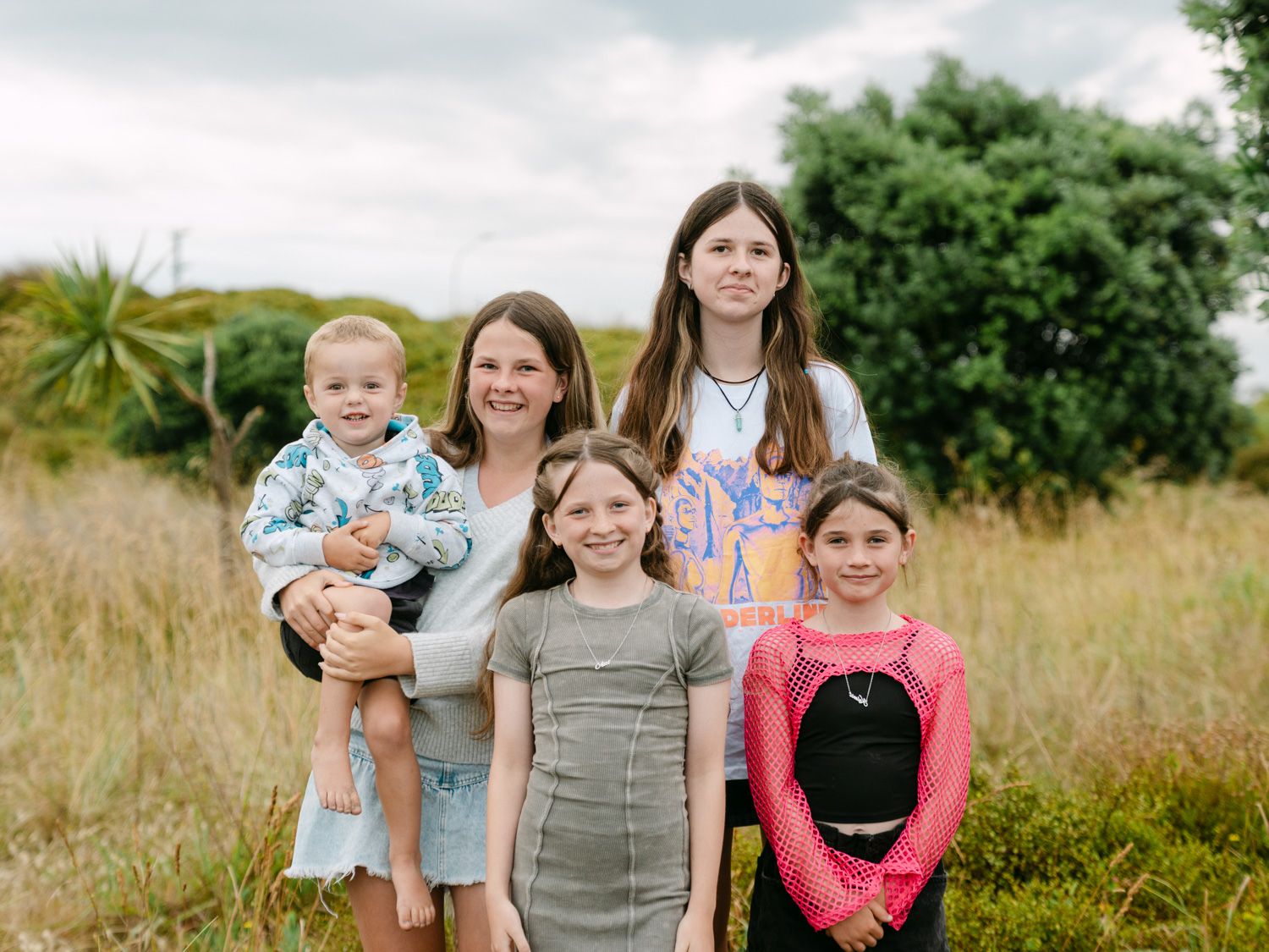 A woman holding a toddler poses outdoors with four children in a grassy field under a cloudy sky.