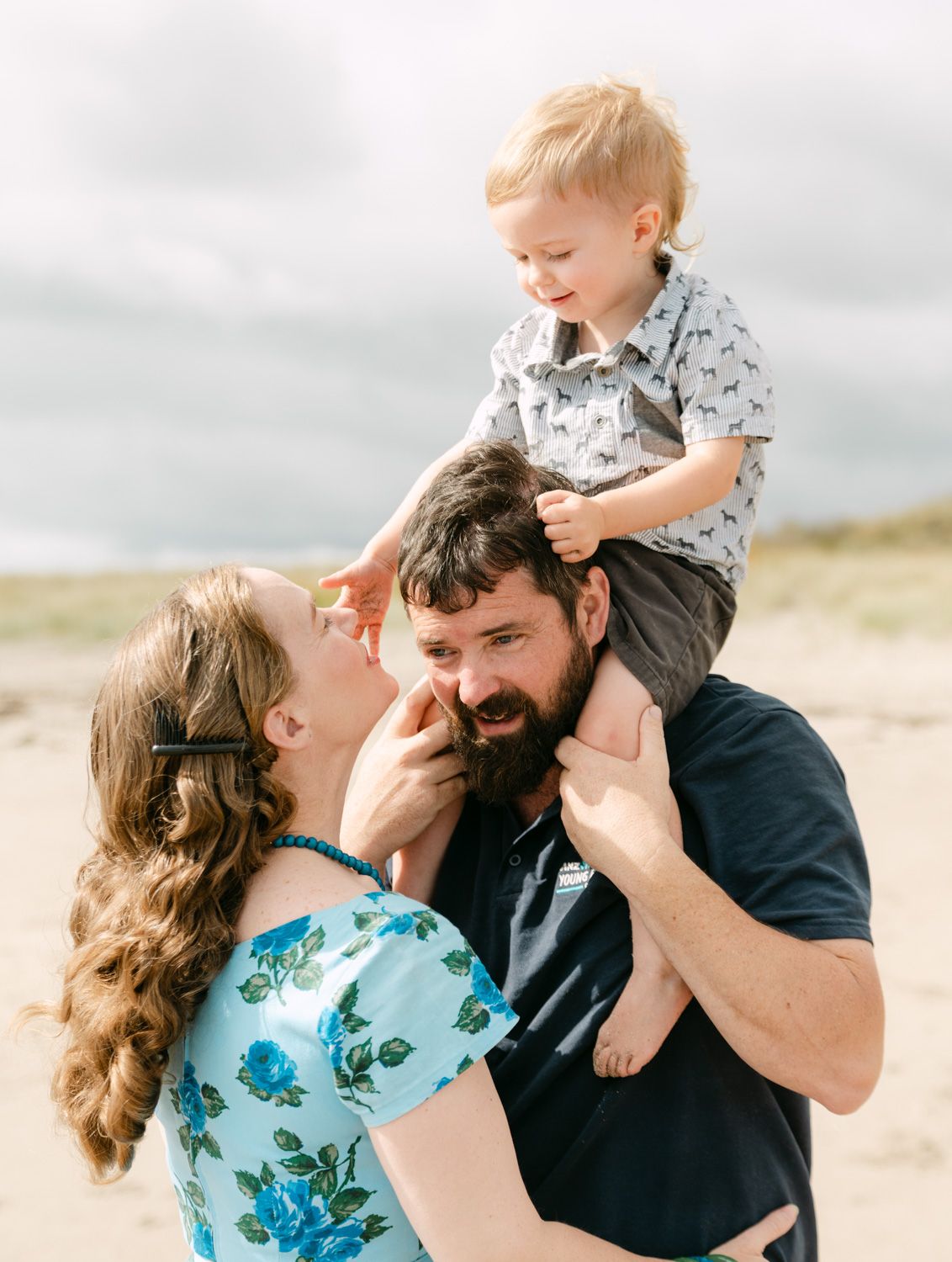 A happy family of three enjoys a beach day, with a toddler sitting on dad's shoulders while mom smiles lovingly.