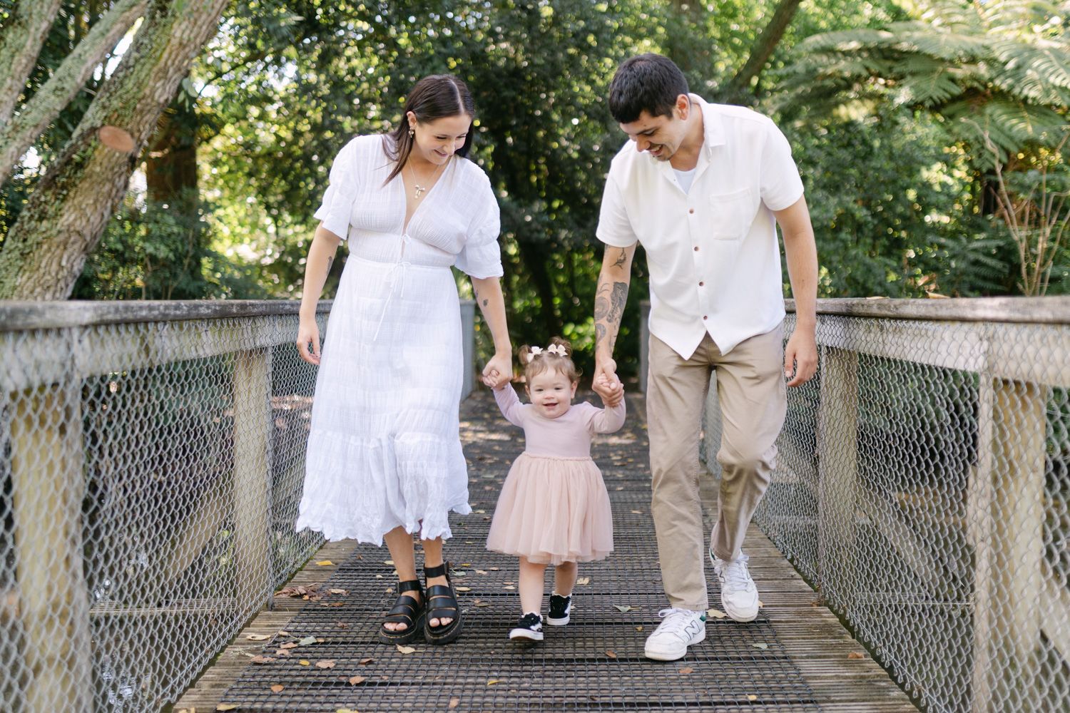 A young family walks hand-in-hand on a wooden bridge, parents guiding their toddler daughter through a lush green park.