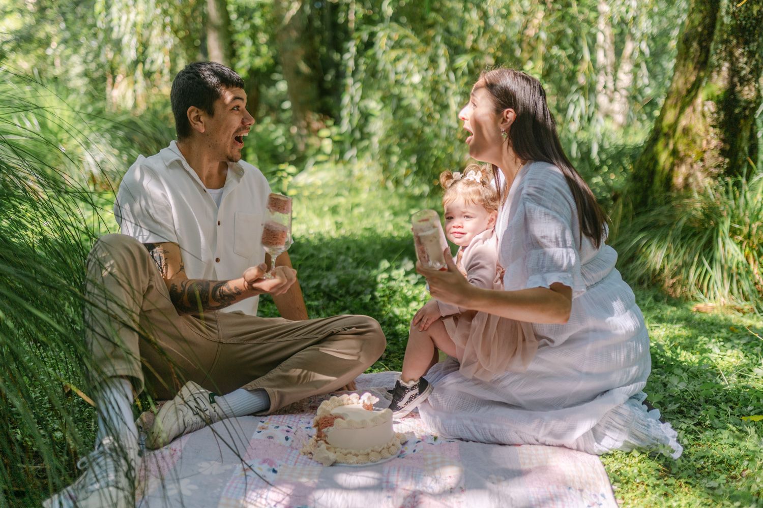 A happy family enjoying a sunny outdoor picnic on a blanket surrounded by lush green trees.