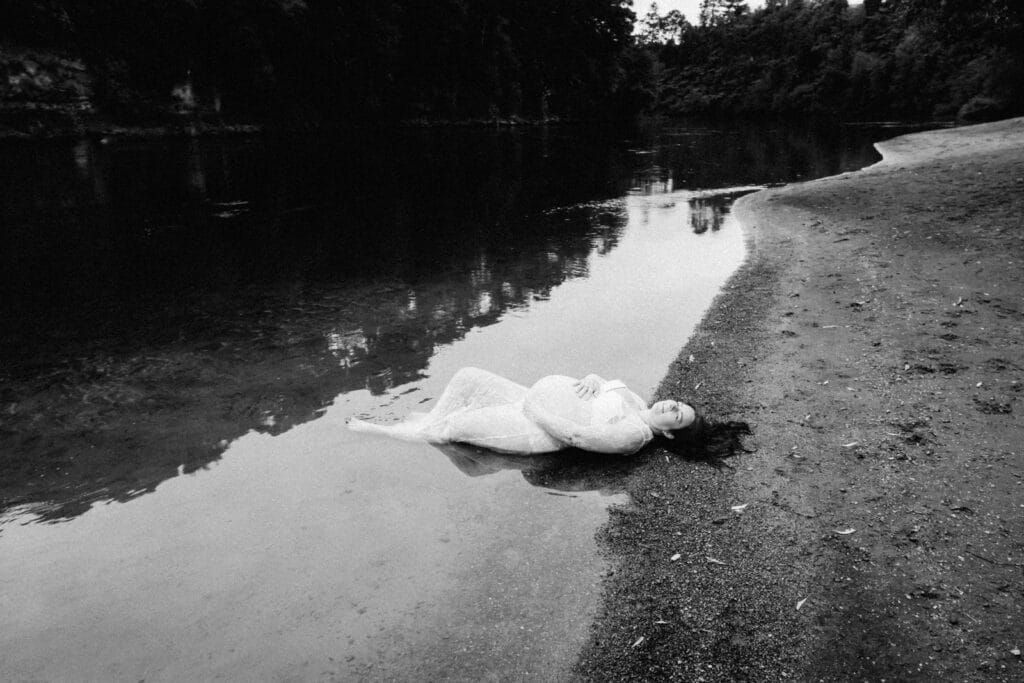 Black and white photo of a person in white dress lying on a sandy riverbank beside calm reflective water.