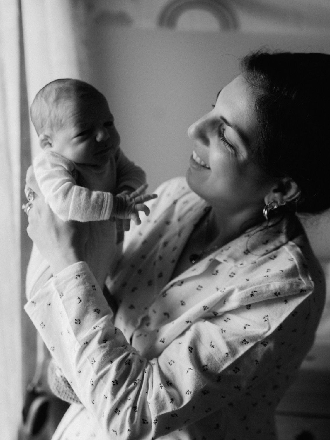 Black and white photo of a smiling mother holding her newborn baby up near a bright window.