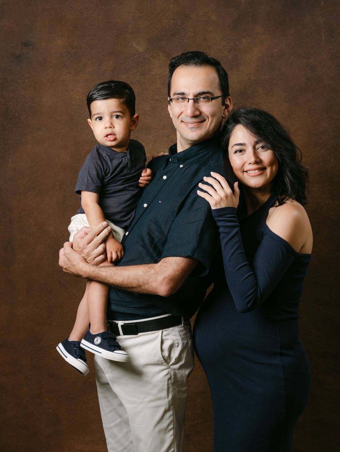 A family portrait of a man holding a toddler boy and a woman in a navy dress posing together in a studio.