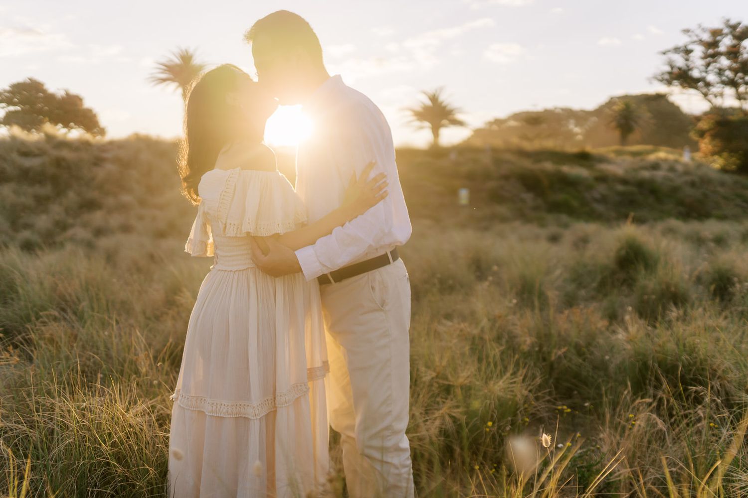 Couple embracing at golden sunset in a grassy field, woman in white dress, warm romantic outdoor photography.