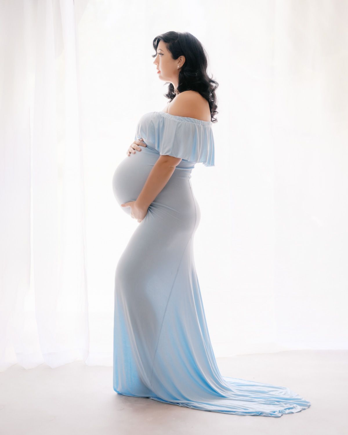 Pregnant woman in elegant light blue off-shoulder maternity gown posing sideways in bright studio setting.