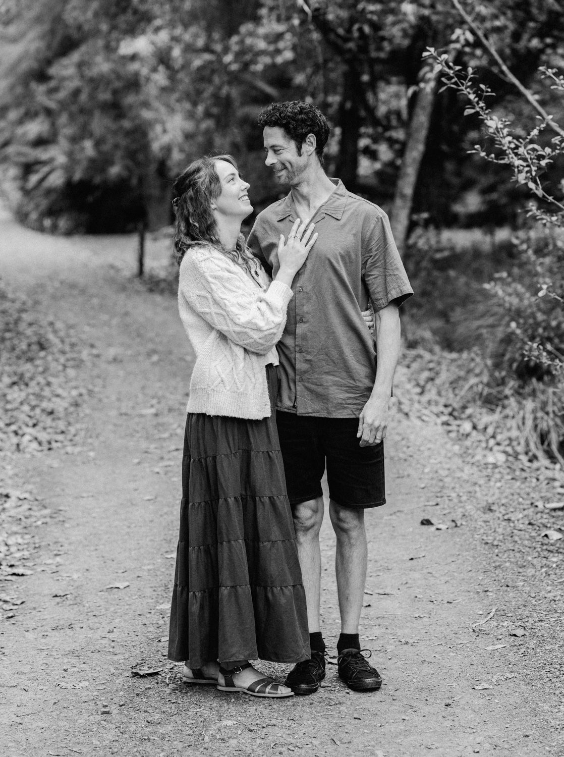 Black and white photo of a smiling couple standing on a wooded path, woman in flowy skirt touching mans chest.