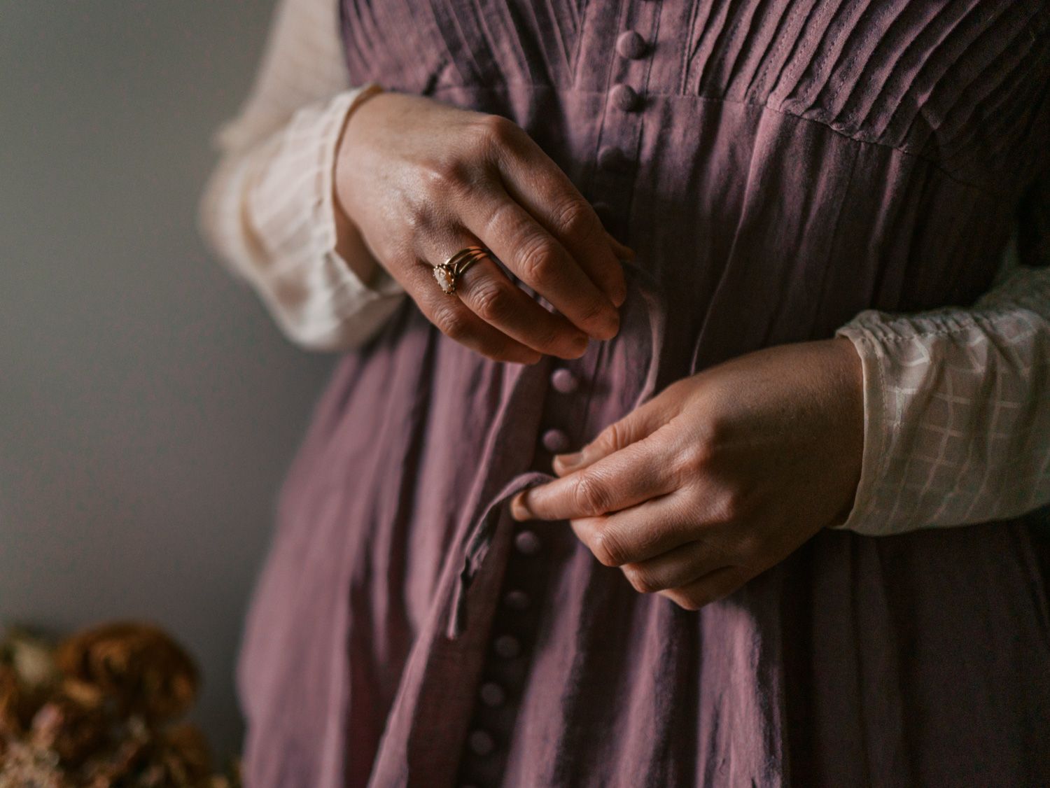 Woman in vintage purple dress fastening buttons, wearing a ring, with white lace sleeves visible.